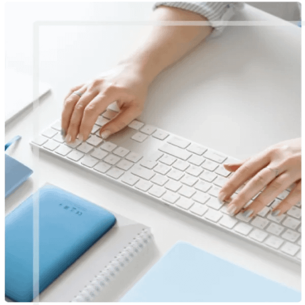 A pair of hands typing on a white keyboard, resting on a clean, white surface. Nearby are a notebook and other blue accessories, suggesting a modern workspace.