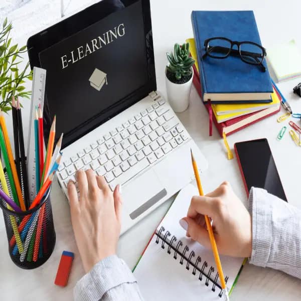 A desk setup with a laptop open to an e-learning program. The desktop is full of books, a notebook and pencil. The laptop displays "E-Learning".

