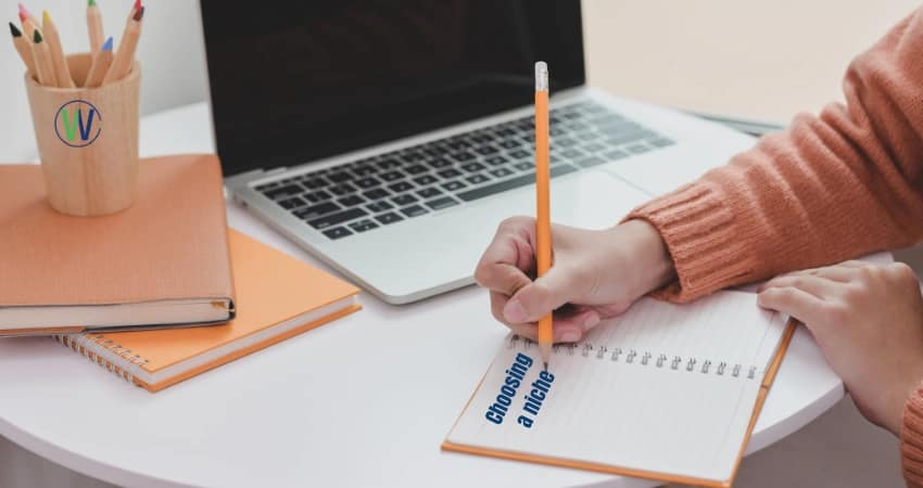 Person writing in a notebook on a desk with a laptop. The notebook has the words "Choosing a niche" written on the cover.