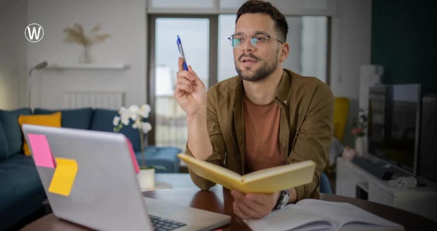 رجل يرتدي نظارات يجلس على مكتب مع حاسوب محمول ودفتر ملاحظات، رافعًا قلمًا كأنه يُثبت وجهة نظر. خلفه، غرفة معيشة بأريكة وديكورات.