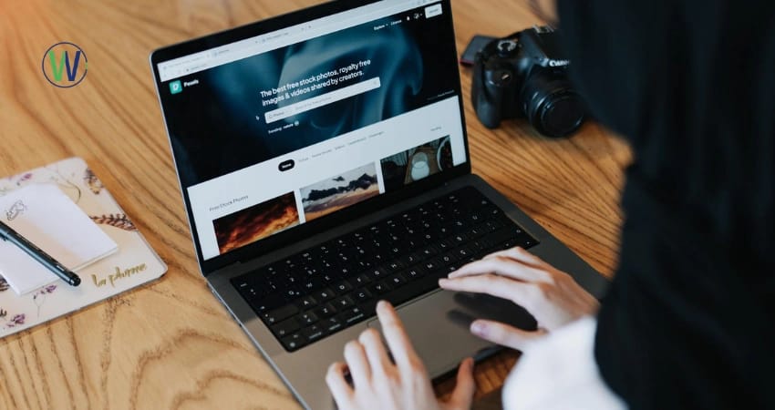 A person's hands are on a computer keyboard and trackpad, there is a camera, and a notebook with a pen next to the laptop. The website on the laptop screen reads: “The best free stock photos, royalty free images & videos shared by creators”.