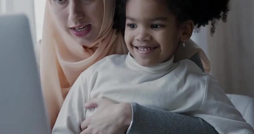 امرأة ترتدي حجابًا بلون الخوخ تحمل طفلًا مبتسمًا في حجرها. يبدو أنهما ينظران إلى جهاز كمبيوتر محمول.