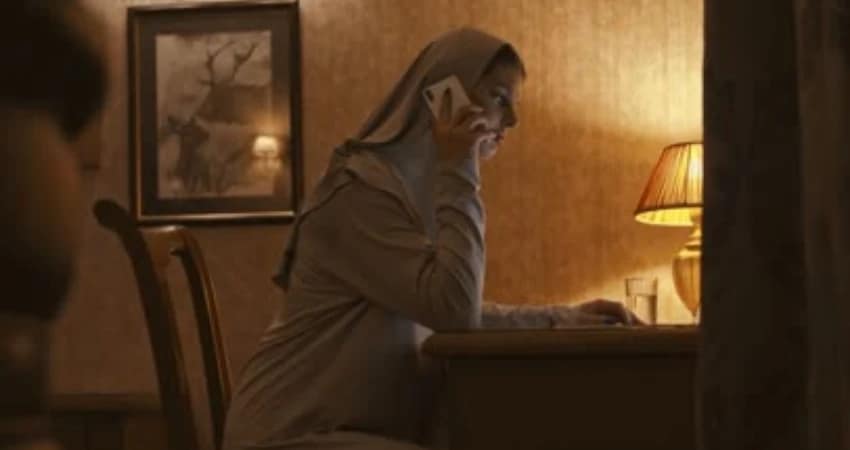 في غرفة ذات إضاءة خافتة، تظهر امرأة محجبة تتحدث عبر الهاتف. تجلس على مكتب قرب مصباح وصورة مؤطرة.