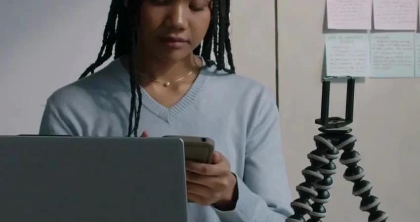 شخص ذو ضفائر، يرتدي قميصًا أزرق، ينظر إلى هاتفه. أمامه حاسوب محمول، وخلفه حامل ثلاثي القوائم وملاحظات لاصقة على الحائط.