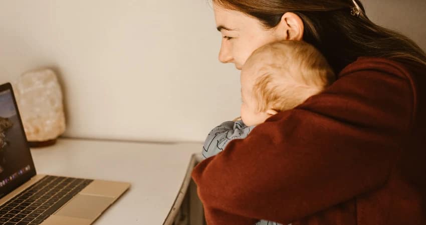 أمٌّ تجلس على مكتبٍ وطفلها بين ذراعيها، تنظر إلى شاشة حاسوبها المحمول. المشهد دافئٌ وجذاب، وبجانب الحاسوب المحمول مصباحٌ ملحيّ.