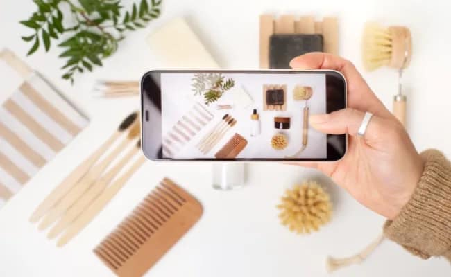 A person takes a cell phone photo of an array of eco-friendly items on a white surface. The items include a comb, brushes, soaps, and a small plant.