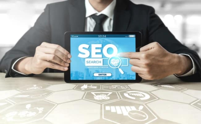 A businessperson holding a tablet displays SEO software. The screen reads “SEO”, “Search” and “Enter”, with a magnifying glass icon. The table has additional SEO icons on it
